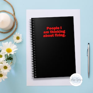 K&ouml;nnte beinhalten: Ein schwarzes Spiralnotizbuch mit dem Text "People I am thinking about firing" in Rot. Das Notizbuch liegt auf einem wei&szlig;en Blatt Papier, mit einem Stift und Blumen im Hintergrund.