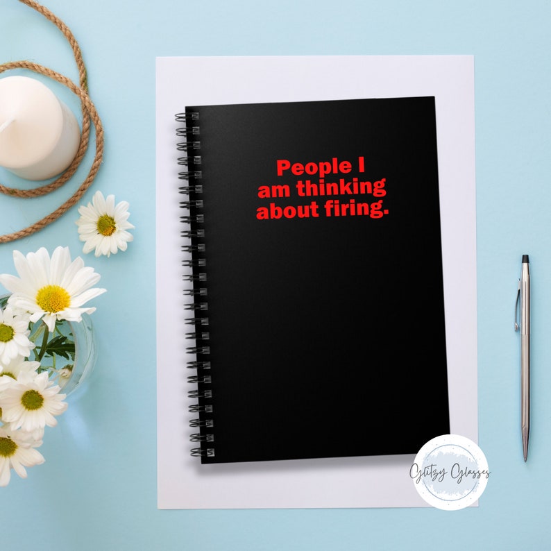 K&ouml;nnte beinhalten: Ein schwarzes Spiralnotizbuch mit dem Text "People I am thinking about firing" in Rot. Das Notizbuch liegt auf einem wei&szlig;en Blatt Papier, mit einem Stift und Blumen im Hintergrund.