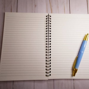 K&ouml;nnte beinhalten: Ein offenes Notizbuch mit linierten Seiten und einer schwarzen Spiralbindung, zusammen mit einem hellblauen Stift mit goldenen Akzenten. Der Stift liegt auf der rechten Seite des offenen Notizbuchs. Der Hintergrund ist eine helle Holzoberfl&auml;che.