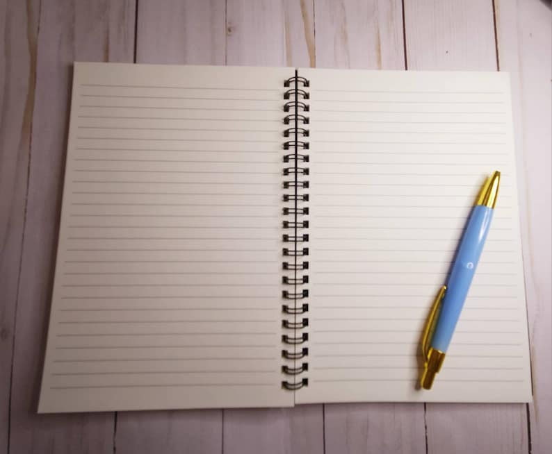 K&ouml;nnte beinhalten: Ein offenes Notizbuch mit linierten Seiten und einer schwarzen Spiralbindung, zusammen mit einem hellblauen Stift mit goldenen Akzenten. Der Stift liegt auf der rechten Seite des offenen Notizbuchs. Der Hintergrund ist eine helle Holzoberfl&auml;che.