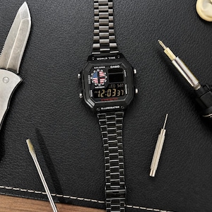 May include: A black Casio digital watch with a metal link band, featuring a World Time display, an American flag graphic on the dial, and the text Illuminator. The watch face shows the time 12:03:37, the date WED 5-22, and 10 Year Battery text.