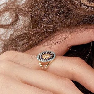 May include: A gold ring featuring a circular face with a central cluster of small, round, light-colored stones surrounded by a concentric border of dark blue stones, set on a thin, polished gold band that splits into two delicate, open-ended arms.