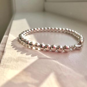 May include: A silver bracelet featuring faceted beads. The bracelet is composed of small, round, shiny beads linked together. The clasp is visible on the right. The bracelet rests on a white surface, with shadows cast across it.