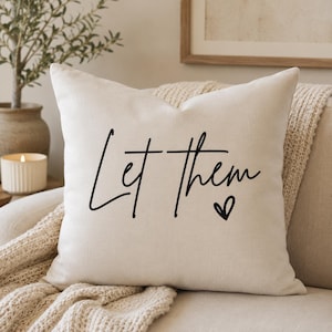Peut inclure: Un coussin carré blanc cassé avec le texte cursif noir Let Them et une petite icône de cœur noir centrée sur le devant. Le coussin présente une texture de tissu lisse et est placé à la verticale contre un coussin de canapé de couleur neutre.