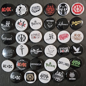 May include: A collection of thirty-six circular pin-back buttons featuring various rock and metal band logos and graphics, including AC/DC, Deftones, Disturbed, Stone Sour, Trivium, and Dream Theater, displayed in a grid on a dark surface.