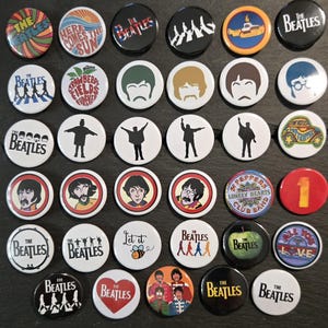May include: A collection of thirty-five circular pin-back badges featuring various designs related to The Beatles, including band member silhouettes, album art, song titles like Here Comes The Sun and Let It Be, and iconic band logos in diverse colours.