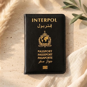 May include: A black passport cover featuring the gold-embossed text INTERPOL, the Arabic translation, a central globe and sword emblem, and the words PASSPORT, PASSEPORT, PASAPORTE, and Arabic text, finished with a small gold rectangular icon.