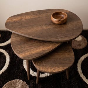 May include: A trio of nesting tables in a dark brown wood grain finish. The tables have an unusual, organic shape with rounded edges and tapered legs. A small wooden bowl sits on the top table. The tables are arranged on a patterned rug.