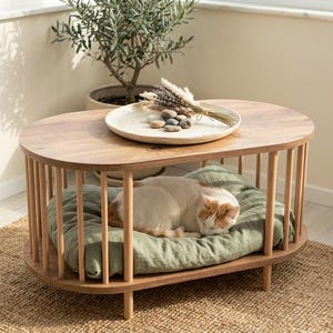 May include: An oval wooden side table with vertical spindle supports and a lower shelf. A white speckled ceramic plate with stones and dried lavender sits on top. A white and ginger cat rests on a sage green cushion on the lower shelf.