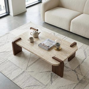 May include: A rectangular coffee table with a beige travertine stone top and dark brown wooden legs. On the table sits a white ceramic teapot, a matching teacup, a book titled Minimalist Interiors, and a small succulent inside a glass cloche.