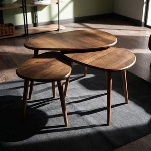 May include: A set of three nesting tables crafted from dark brown wood with a visible grain. Each table features a smooth, rounded, irregular teardrop-shaped top supported by three tapered wooden legs, arranged in a tiered, overlapping formation.