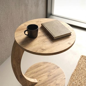May include: A round wooden side table with a C-shaped base and a natural wood grain finish. A matte black ceramic mug and a closed rectangular book with a textured beige cover are placed on the circular tabletop surface of the furniture piece.