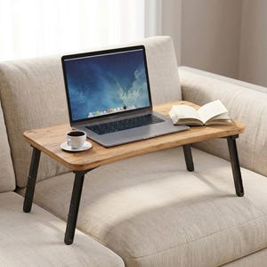 May include: A portable wooden laptop desk with four black foldable legs. The rectangular tabletop features a natural wood grain finish. A silver laptop, a white ceramic coffee cup on a matching saucer, and an open book are placed on the surface.
