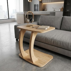 Può includere: Un tavolino a forma di C in legno chiaro con un piano rettangolare piatto e una base di supporto curva. Sul piano del tavolo si trovano un supporto per cuffie in metallo dorato con cuffie, una penna argentata e un taccuino chiuso.