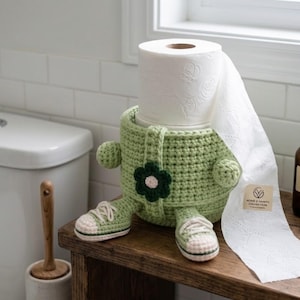 May include: A light green crocheted toilet paper holder shaped like a character with small arms, legs, and sneakers. A dark green flower is attached to the front. A roll of white toilet paper sits inside, with a label reading "KONG'S VANITY COLLECTION".