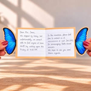 Può includere: Un biglietto rettangolare con bordo arancione chiaro e ali di farfalla blu brillante attaccate su entrambi i lati. Il biglietto presenta un testo scritto a mano indirizzato alla Sig.ra Jones, con una visita, un orario e JW.ORG.