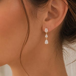 Peut inclure: Une boucle d'oreille pendante en or avec trois pierres transparentes alignées verticalement. La pierre supérieure est ronde, celle du milieu est en forme de poire et la pierre inférieure est une goutte plus grande en forme de poire.