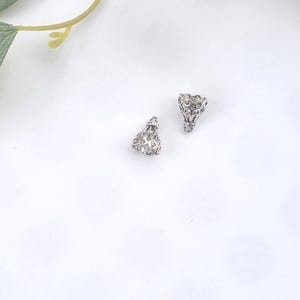 May include: Two silver-toned metal bead caps featuring an intricate, openwork filigree design. Each piece has a bell-like shape with a small loop at the top, showcasing a detailed, textured pattern throughout the surface of the metal components.