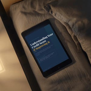 May include: A black tablet displaying the text "Understanding Your Credit Score & Improving It" on a dark blue background. The tablet is resting on a gray pillow and comforter, with a wooden surface visible in the background.