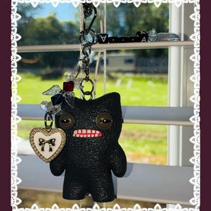 May include: A black plush doll keyring with a heart-shaped charm. The doll has large, round eyes and a stitched mouth with visible teeth. The heart charm is white with a gold bow. The keyring is hanging in front of a window.