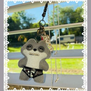 May include: A grey plush sloth keyring wearing camouflage patterned underwear. The sloth has a stitched mouth and button eyes. The keyring includes a metal clasp and charms. Text at the top reads "+ Free Stickers" and at the bottom "+ Free Cards".