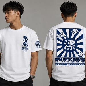 May include: A white short-sleeve t-shirt featuring blue graphic prints. The front displays Japanese characters, the text AKUMA MOTOR DIVISION EST. 2020, and a sleeve logo. The back features a large sunburst design with clouds and RPM OPTIC GARAGE text.