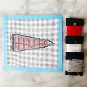 May include: A square needlepoint canvas featuring a gray pennant design with the word HARVARD stitched in red, bordered by light blue tape. Below the pennant is a small pink heart and the letters CC. Three spools of embroidery thread are included.