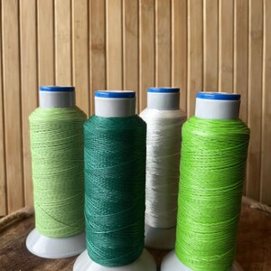 May include: Four spools of thread in shades of green and white. The spools are on white bases with blue and white tops. The thread colors include light green, dark green, and white, with a bright green spool on the right.