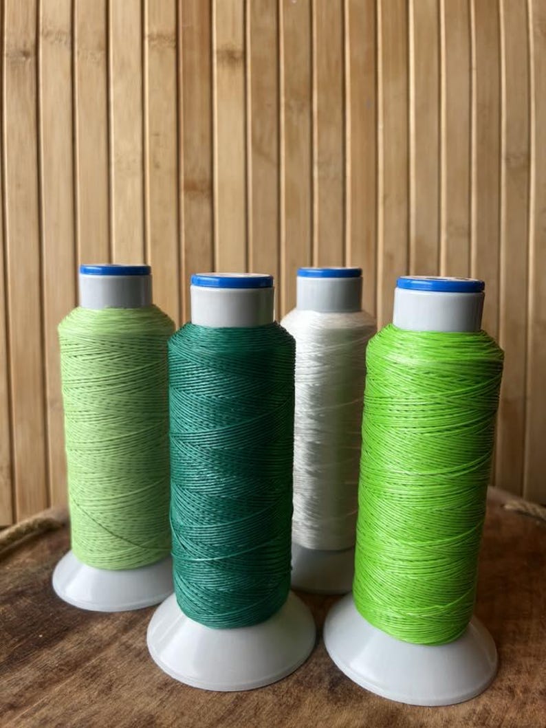 May include: Four spools of thread in shades of green and white. The spools are on white bases with blue and white tops. The thread colors include light green, dark green, and white, with a bright green spool on the right.