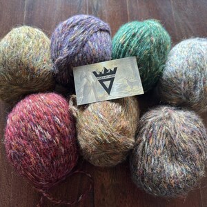 Peut inclure: Sept boules de fil coloré dans les tons de vert, violet, rouge et marron. Une petite carte avec un logo est placée sur le dessus de certaines boules de fil. Le fil est disposé sur une surface en bois foncé.