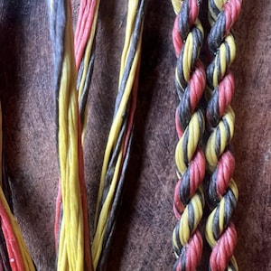 Pode incluir: Close-up de cordões coloridos e cordas trançadas. Os cordões são vermelhos, amarelos e pretos. As cordas trançadas apresentam um padrão torcido de vermelho, amarelo e preto. O fundo é uma superfície texturizada marrom escuro.