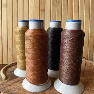 May include: Four spools of thread in various colors: gold, orange, black, and brown. Each spool is on a white base with a blue top. The background is a light brown bamboo wall and a wooden surface.
