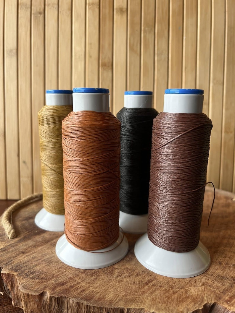 May include: Four spools of thread in various colors: gold, orange, black, and brown. Each spool is on a white base with a blue top. The background is a light brown bamboo wall and a wooden surface.