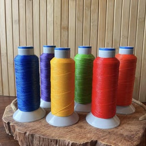 May include: Six spools of colorful thread in blue, purple, yellow, green, red, and orange. Each spool is on a white base with a blue top. The spools are arranged on a wooden slab with a rope handle.