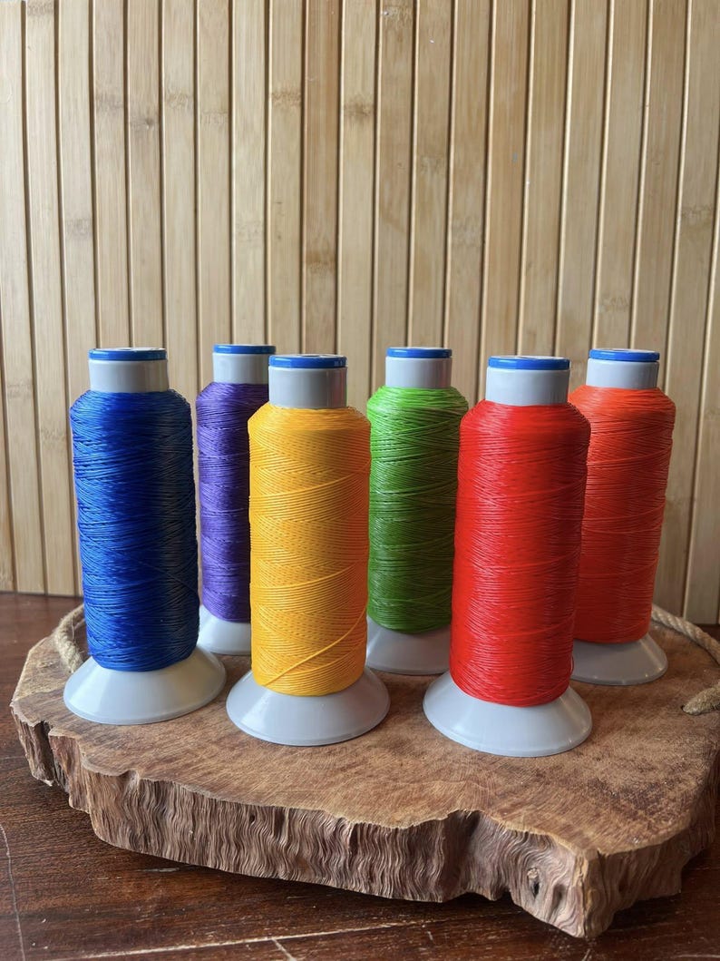 May include: Six spools of colorful thread in blue, purple, yellow, green, red, and orange. Each spool is on a white base with a blue top. The spools are arranged on a wooden slab with a rope handle.