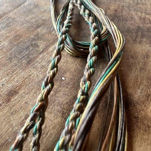 May include: Close-up of a collection of braided cords in shades of brown, green, and yellow. The cords are intertwined, creating a textured and layered effect. The image highlights the intricate braiding and the natural color palette of the cords.