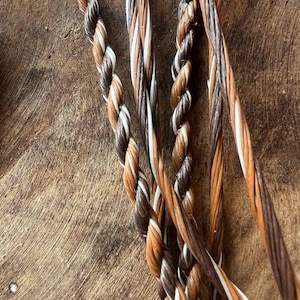 May include: Close-up of several strands of braided cord in shades of brown, white, and tan. The cords are tightly twisted together, creating a textured appearance. The background is a wooden surface.