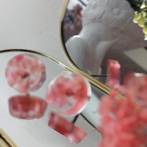 May include: A pair of round stud earrings featuring a clear resin base with embedded pink floral elements. The earrings are positioned on a reflective surface, showing their circular shape and the pink botanical details captured within the transparent material.