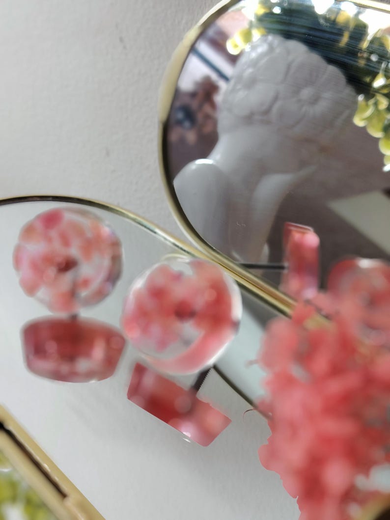 May include: A pair of round stud earrings featuring a clear resin base with embedded pink floral elements. The earrings are positioned on a reflective surface, showing their circular shape and the pink botanical details captured within the transparent material.