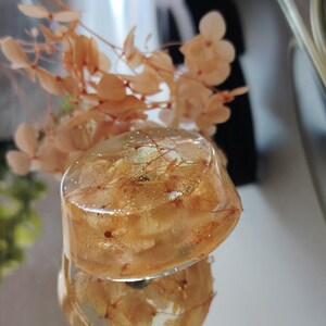May include: A circular, clear resin paperweight or decorative object containing dried, light orange hydrangea petals and fine gold glitter suspended within the material. The object has a smooth, polished surface and a rounded, dome-like shape.