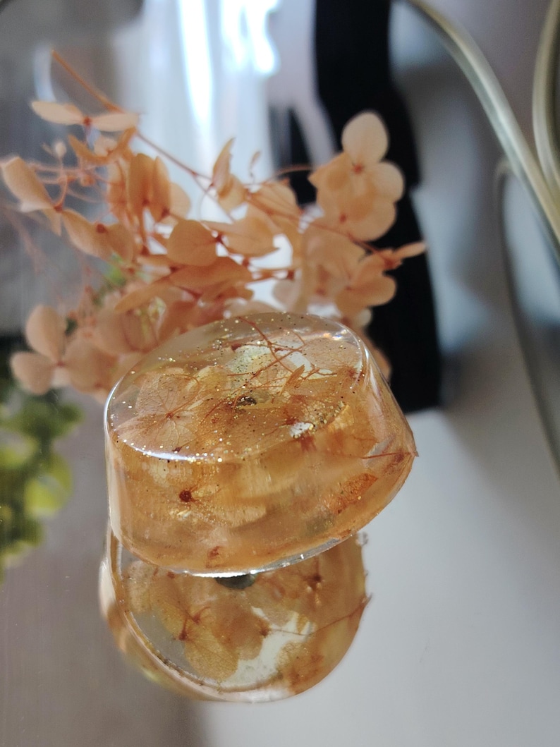May include: A circular, clear resin paperweight or decorative object containing dried, light orange hydrangea petals and fine gold glitter suspended within the material. The object has a smooth, polished surface and a rounded, dome-like shape.