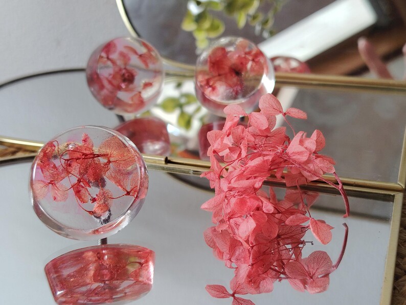 May include: Three circular resin knobs featuring embedded dried pink hydrangea petals. The clear, polished resin discs showcase the delicate floral structure within. Each knob is mounted on a metal base, reflecting the vibrant pink blossoms.