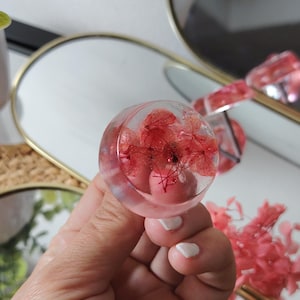 May include: A circular, clear resin ring featuring embedded red hydrangea petals. The transparent material showcases the delicate, natural structure of the dried flowers within the rounded, smooth surface of the jewellery piece.