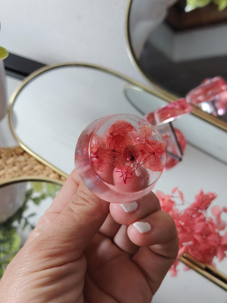 May include: A circular, clear resin ring featuring embedded red hydrangea petals. The transparent material showcases the delicate, natural structure of the dried flowers within the rounded, smooth surface of the jewellery piece.