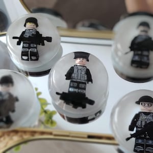 May include: Five circular clear resin plugs, each containing a miniature plastic soldier figure wearing a black helmet, tactical vest, and carrying a rifle. The figures are embedded within the transparent material, displaying detailed black and gray uniforms.