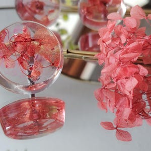 May include: A round, clear resin cabinet knob featuring embedded pink hydrangea petals. The knob has a smooth, glossy finish and a metallic base. A cluster of loose, dried pink hydrangea flowers is positioned next to the knob.