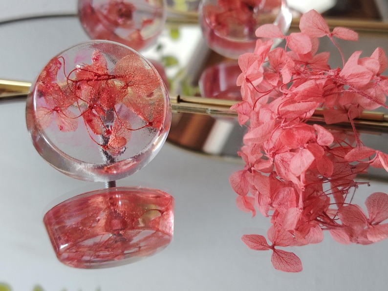 May include: A round, clear resin cabinet knob featuring embedded pink hydrangea petals. The knob has a smooth, glossy finish and a metallic base. A cluster of loose, dried pink hydrangea flowers is positioned next to the knob.