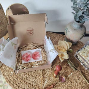 May include: A brown cardboard gift box containing two circular resin pieces with embedded pink floral elements, nestled in shredded paper filler. The box lid interior features a circular sticker with the text Handmade with Love.