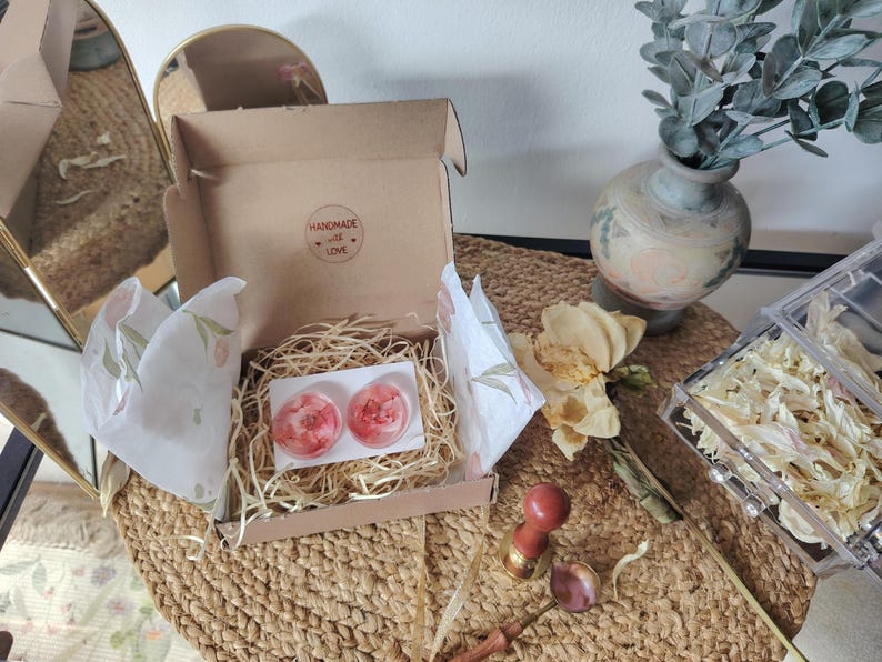 May include: A brown cardboard gift box containing two circular resin pieces with embedded pink floral elements, nestled in shredded paper filler. The box lid interior features a circular sticker with the text Handmade with Love.
