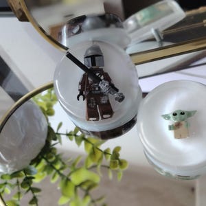 May include: Two clear circular resin ornaments featuring embedded miniature figures. One contains a brown and silver armored character with a helmet and rifle, while the other contains a small green creature with large ears and a tan robe.